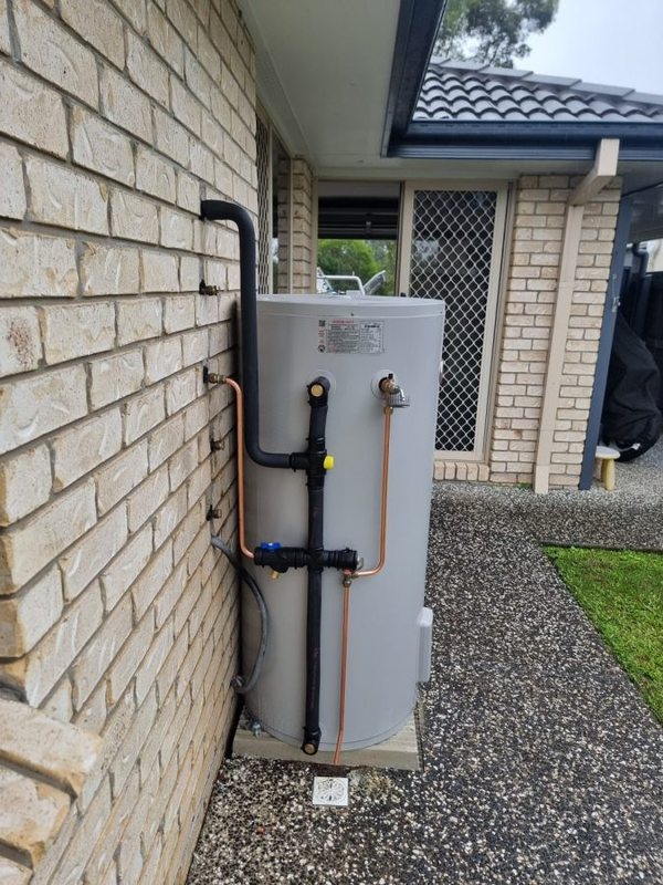 Electric hot water storage unit against a brick wall, with copper and black pipework